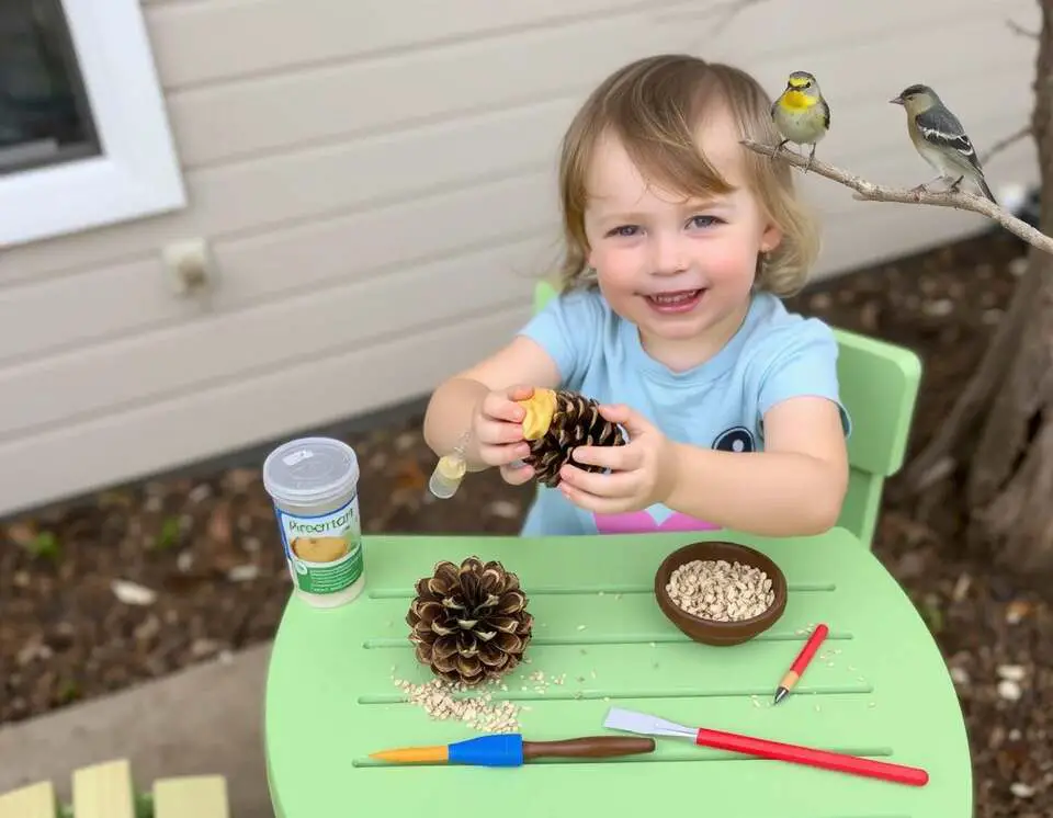 A preschooler making a pinecone bird feeder with peanut butter and seeds at an outdoor table, with birds nearby.